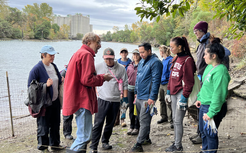 A group of people stand and listen during a shoreline restoration activity beside a river. One person in the foreground speaks while others gather around holding gloves or tools. The setting includes a dirt path, a wire fence, overhanging tree branches, and industrial buildings across the water under an overcast sky.