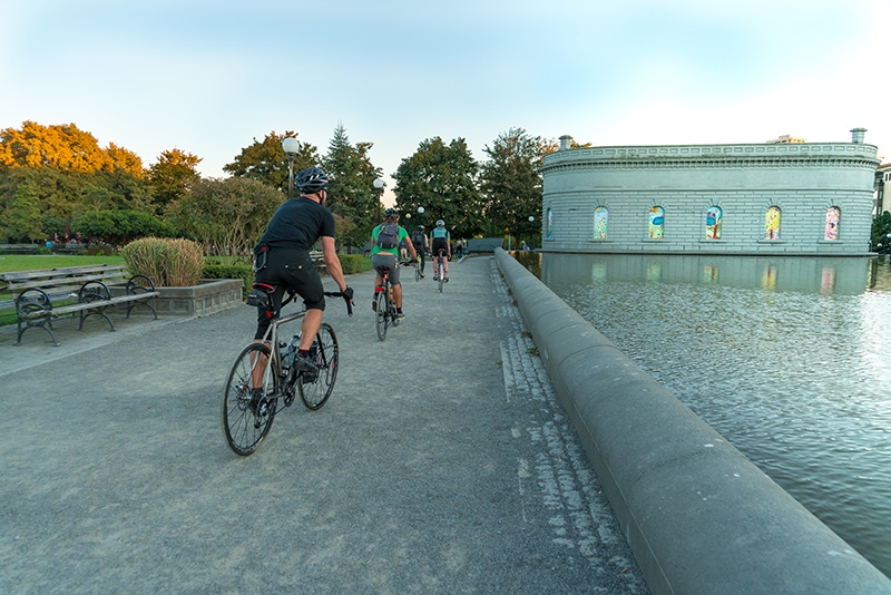 Cyclists at Cal Anderson Park riding along a pathway beside the reflecting pool, with trees and a round stone building in the background.
