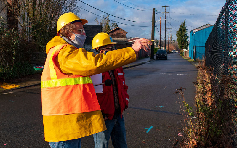 Two people wearing hard hats, face coverings, and high‑visibility safety gear stand on a neighborhood street beside a chain‑link fence. One person points toward the fence while the other looks on. The scene includes utility poles, overhead wires, houses, and a blue industrial building under partly cloudy sky.