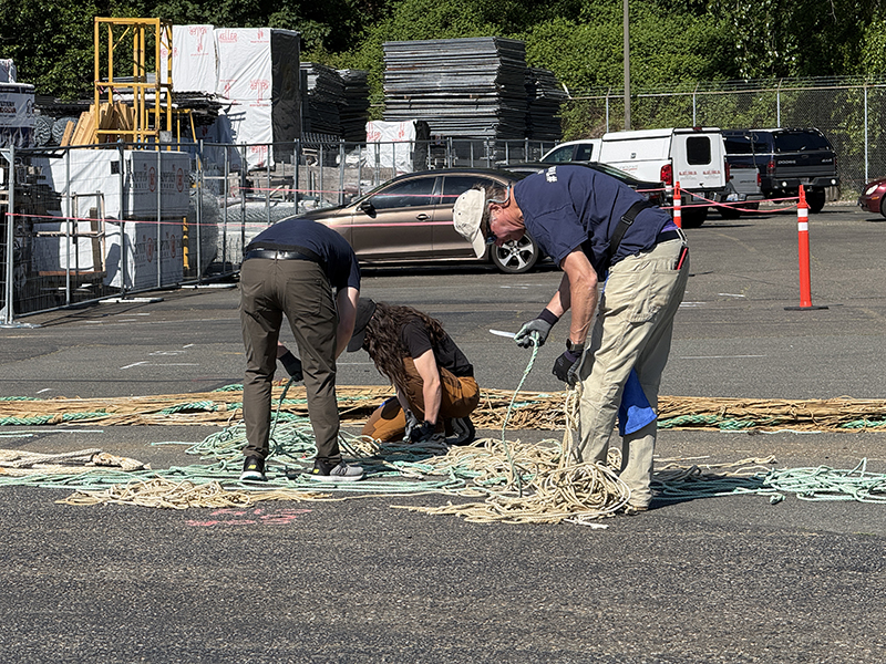 Three people are working together in an outdoor lot, sorting and coiling long bundles of light‑colored rope spread across the pavement. Construction materials, fencing, parked vehicles, and trees are visible in the background.