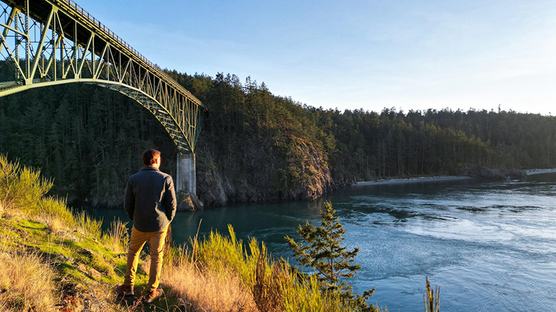 A person stands on a grassy hillside under the Deception Pass Bridge, looking out over the water and the forested shoreline.