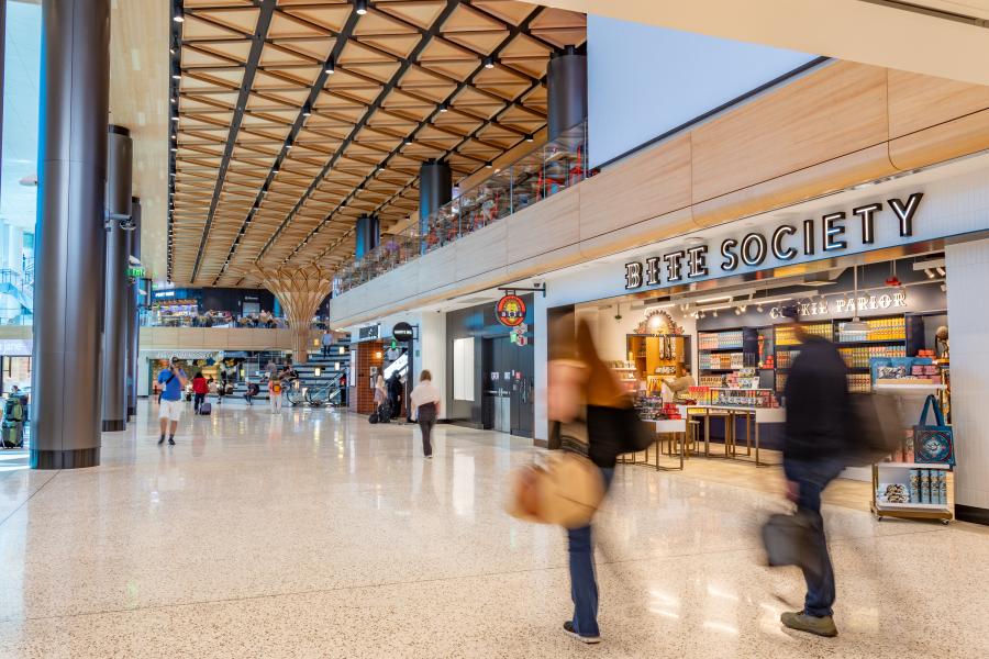 A spacious airport concourse with travelers walking past a shop called “Bite Society,” featuring a high wood‑patterned ceiling and polished terrazzo floors.
