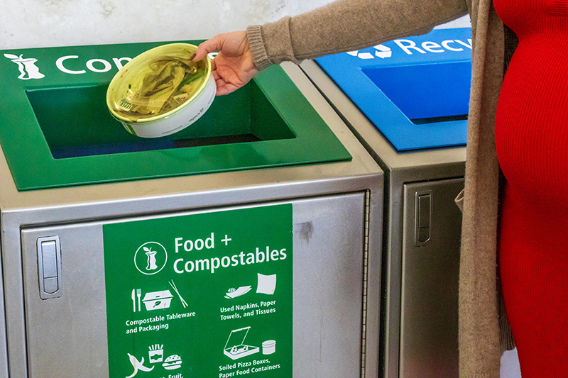 A person places a compostable food container into a green bin labeled “Food + Compostables,” next to a blue recycling bin.
