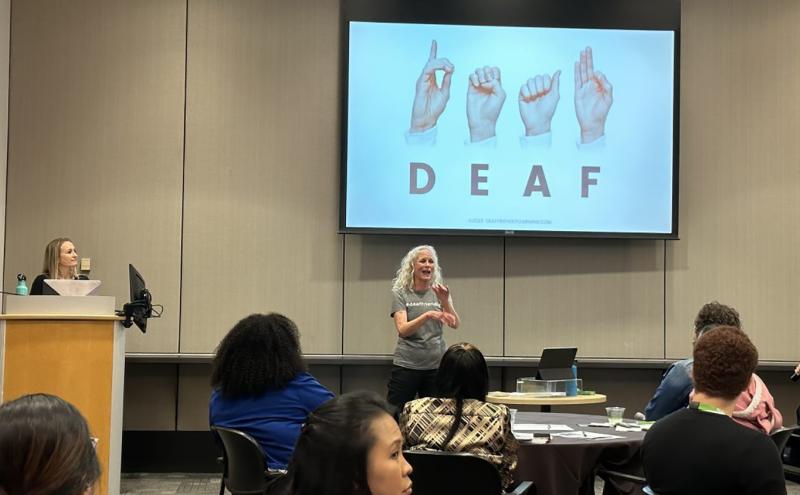 A training room with attendees seated at tables facing the front, where one presenter stands and communicates using hand and arm motions. Another individual stands at a podium on the left. A large screen at the front displays four illustrated hands forming letters in American Sign Language that spell the word “DEAF.”