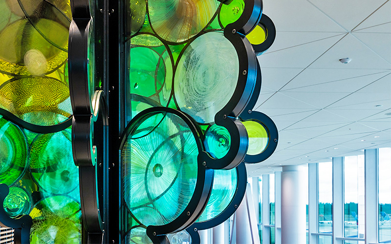 A close‑up view of artist Fumi Amano’s sculpture “Shared Breath,” showing layered circular forms made of translucent green, blue, and yellow materials framed by dark curving structural elements. The textured surfaces and overlapping shapes create a detailed pattern, with the bright interior architecture of the C Concourse Expansion visible in the background.