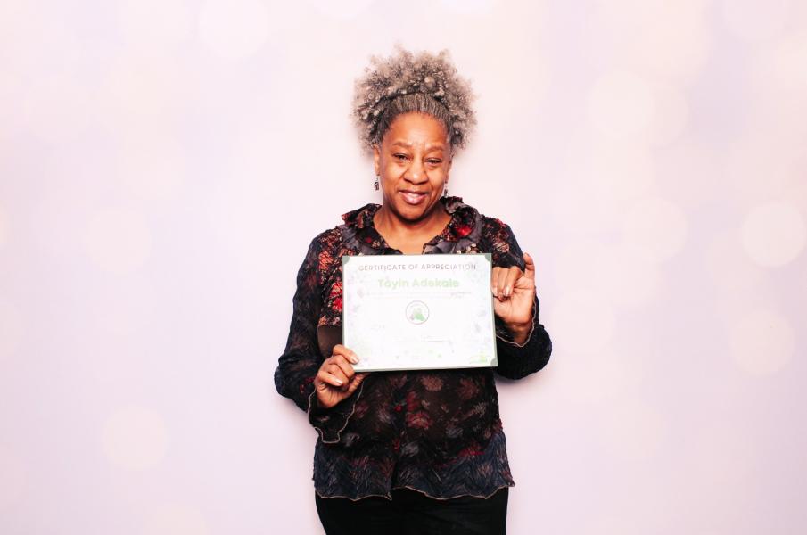 Toyin Adekale standing in front of a light backdrop holding a Certificate of Appreciation with both hands.