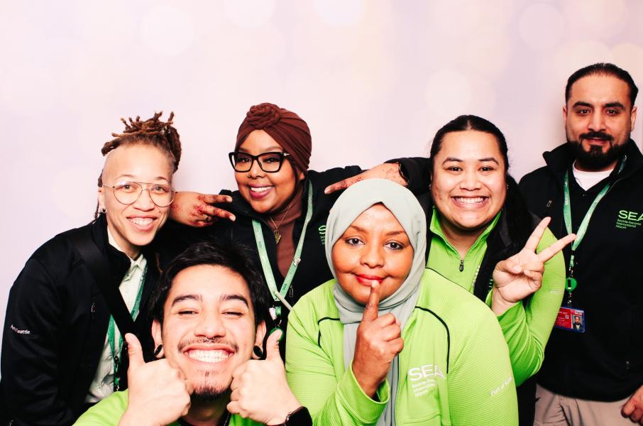 Six people wearing SEA‑branded clothing pose together in front of a light backdrop, making playful hand gestures.