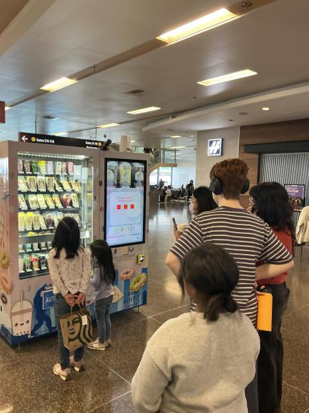 A group of people is gathered in front of a brightly lit vending machine in an indoor public space. The vending machine has two sections: a display window filled with packaged drinks and snacks, and a touchscreen panel showing colorful icons. The people are standing at varying distances, some looking at the touchscreen and others observing the items inside the machine. The background includes seating areas, signage, and overhead lighting within the large indoor area.
