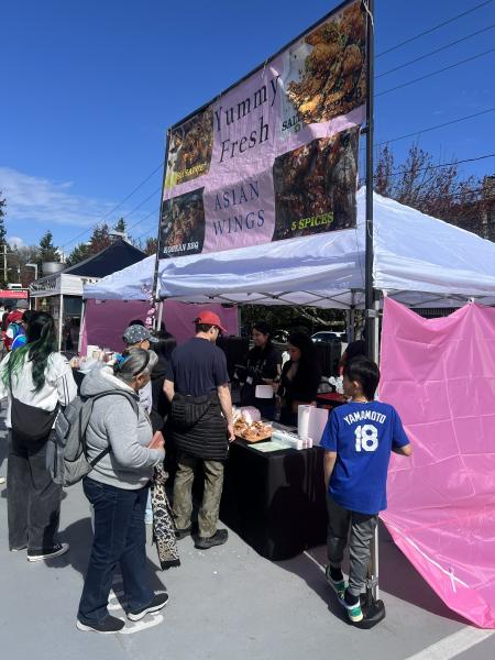 An outdoor food‑booth scene at a market or festival. Several people are standing in front of a vendor tent with a large sign reading “Yummy Fresh Asian Wings” and additional smaller text advertising flavors such as “Korean BBQ” and “5 Spices.” The tent is white with pink side panels, and the vendor inside is serving food from trays on a black table. The area is brightly lit with clear skies, and more tents, people, and trees are visible in the background.