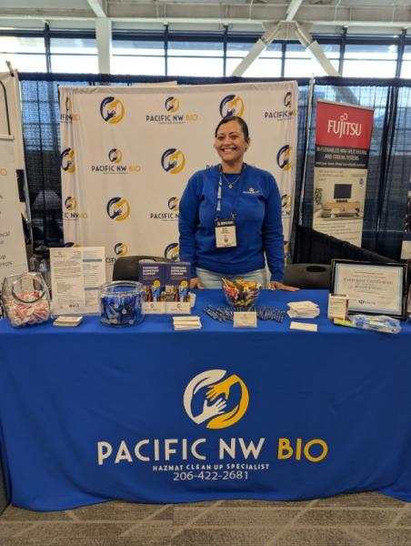 An individual stands behind a table with promotional materials for Pacific NW Bio at an event booth.