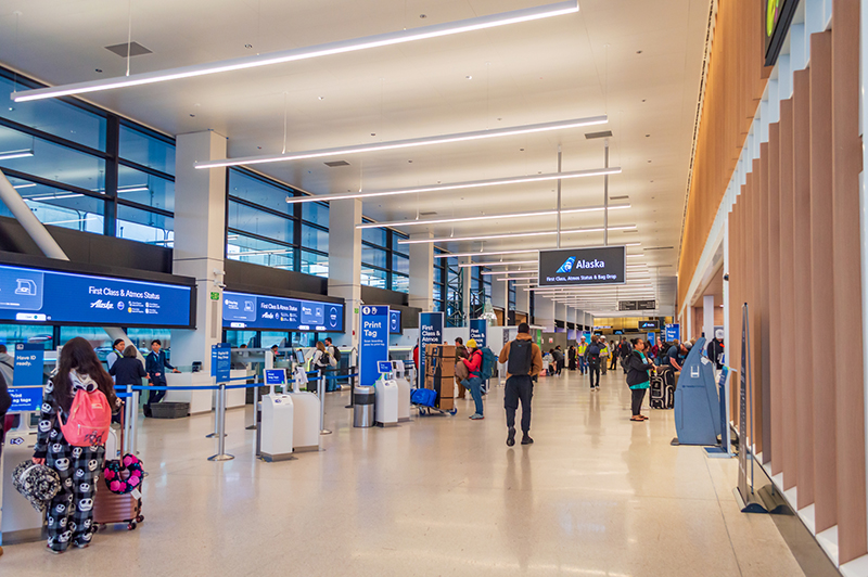 Travelers move through Alaska Airlines’ check‑in area, which features self‑service kiosks, service counters, and overhead digital signs as part of the SEA Gateway project.