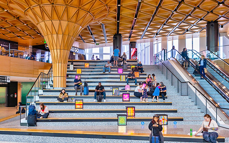 Travelers sit and rest on the Grand Stairs in the C Concourse Expansion. Several illuminated art boxes with colorful, patterned designs are placed along the steps. The space features a tall sculptural column supporting a geometric wood ceiling, large windows, and escalators on both sides. People are seated at different levels of the stairs with carry‑on bags and personal items, creating a busy and open public area.