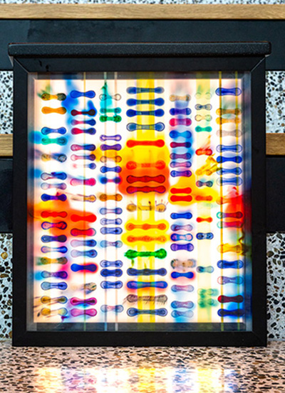 A close‑up view of the illuminated artwork “Surface Tensions,” showing a square lightbox framed in black and filled with rows of colorful abstract shapes in blue, red, yellow, green, and purple against a glowing background.