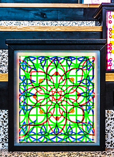 A close‑up view of the artwork “Surface Tensions,” showing a square illuminated panel framed in black. The piece features an intricate geometric pattern made of overlapping red, green, and blue lines that form symmetrical shapes across a bright white background.