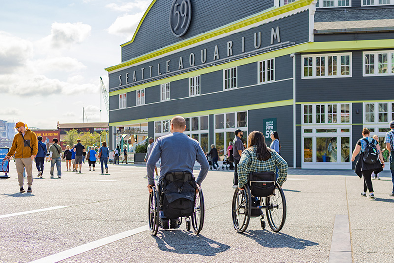 Two people using wheelchairs move along the waterfront in front of the Seattle Aquarium building, with other visitors walking nearby.
