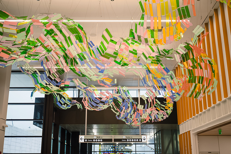New artwork hangs from the ceiling as part of the SEA Gateway project, featuring flowing arrangements of colorful panels suspended above an airport walkway.