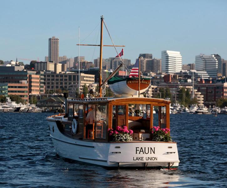 A classic white wooden motorboat named Faun travels on blue water with a city skyline in the background. The boat has varnished wood trim, pink flowers arranged at the stern, and a small white tender secured on the roof. Several flags are displayed, including one with red and white stripes at the back. The shoreline behind the boat is lined with buildings and additional moored vessels.