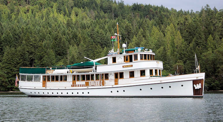 A large classic white motor yacht named Malibu floats on calm water in front of a densely forested shoreline. The yacht features green trim, multiple decks with wooden accents, a mast displaying flags including a Canadian flag, and an emblem of two white swans on the bow.