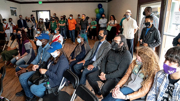 A group of people seated and standing indoors, all wearing face masks, watching an event or presentation.
