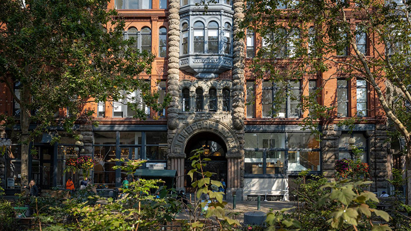 Historic brick building in Pioneer Square with a detailed stone archway and large windows, partly framed by trees in the foreground.