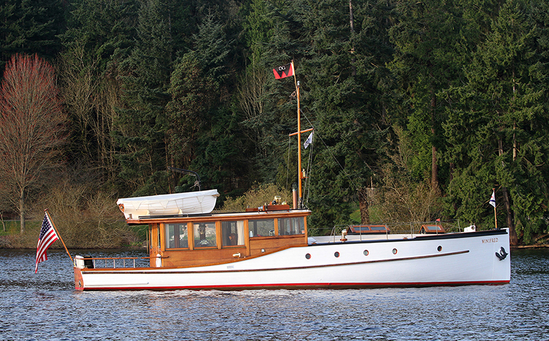 A classic white wooden motorboat named Winifred travels on calm water with a forested shoreline in the background. The vessel features warm wood trim, a covered cabin area, a small white tender stored on the roof, and several flags on its mast, including one near the stern. An American flag flies at the back of the boat.