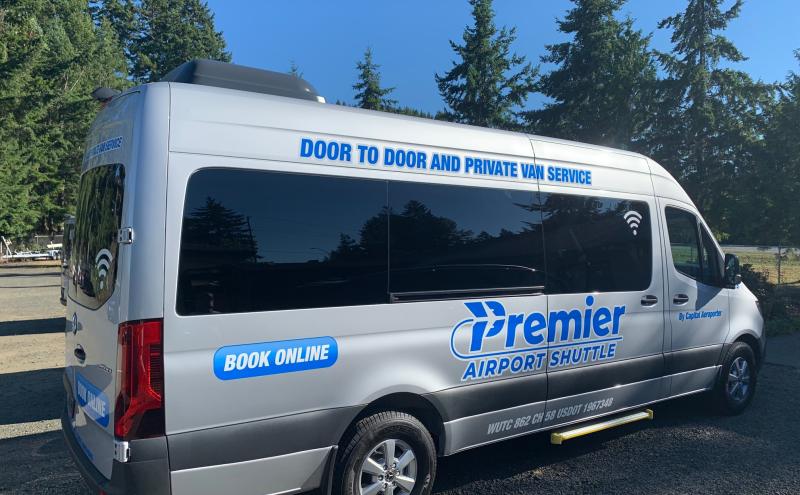 Silver Premier Airport Shuttle van parked outdoors, surrounded by tall evergreen trees. The van displays text including ‘Door to Door and Private Van Service,’ ‘Premier Airport Shuttle,’ and ‘Book Online,’ with the Premier logo on the side.