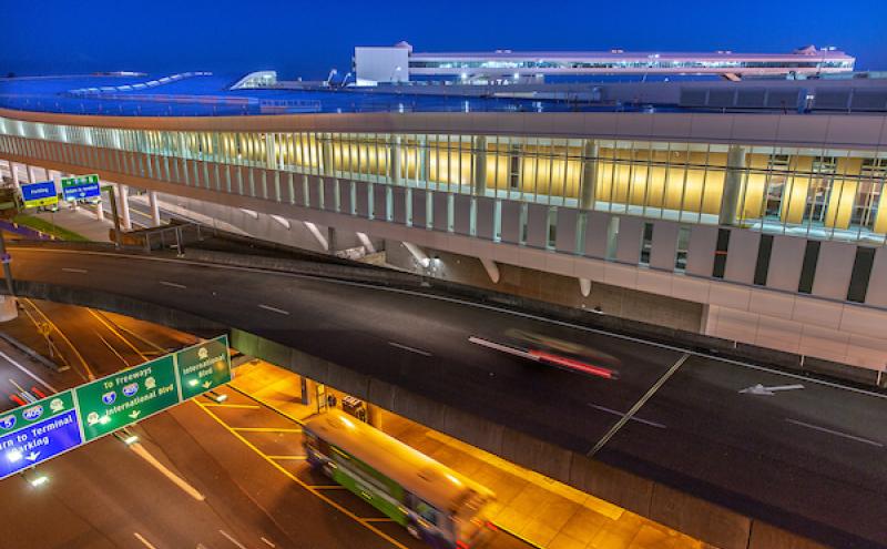 Elevated airport roadway at night with blurred headlights and taillights from moving vehicles beneath a long, modern terminal building with illuminated windows. Overhead signs mark directions to arrivals, the terminal, and parking.
