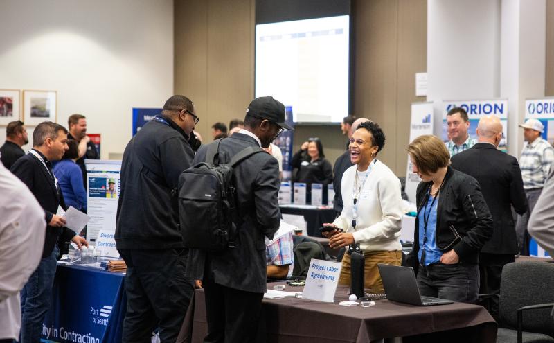 People gather and talk around tables at an indoor event, including a table labeled “Project Labor Agreements” with a laptop and materials. Signs for “Diversity in Contracting” and “ORION” are visible in the background.