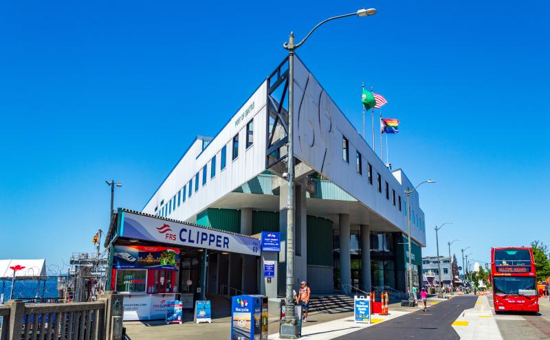 Side angle view of the Port of Seattle’s Headquarters Building at Pier 69, showing the FRS Clipper entrance, street signage, recycling bins, and a red double‑decker tour bus parked along the roadway.