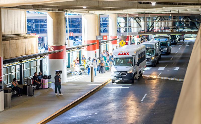 Airport curbside pickup area with shuttle vans lined up, passengers waiting with luggage, and signs marking different pickup zones under a covered roadway.