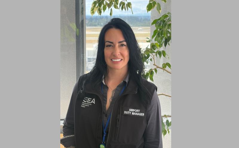 Rachel Sanders, an airport duty manager wearing a SEA Seattle‑Tacoma International Airport vest, stands indoors near a window with the airfield visible in the background and a leafy plant to the side.