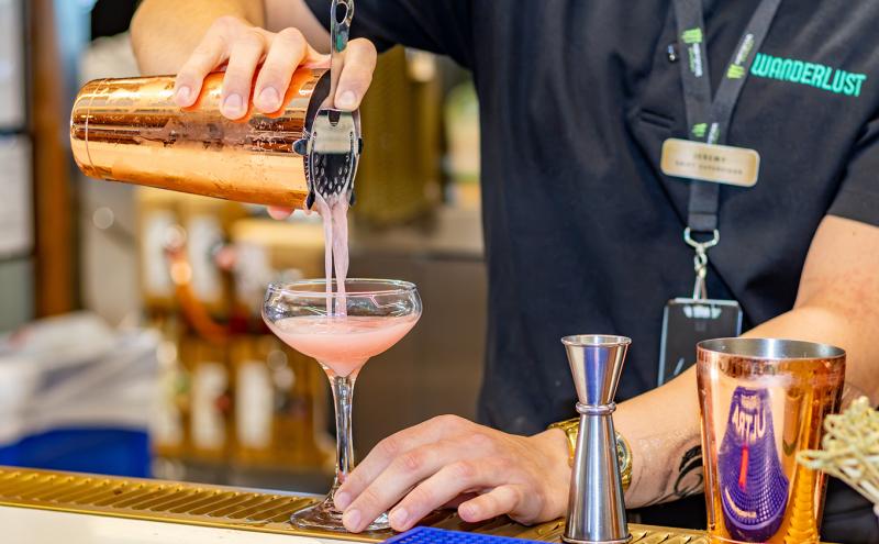 A person wearing a dark shirt with a visible name badge is pouring a pink beverage from a copper-colored cocktail shaker through a strainer into a coupe glass on a bar counter. A metal jigger and another copper-colored shaker are on the counter, with blurred bar shelves and equipment in the background.