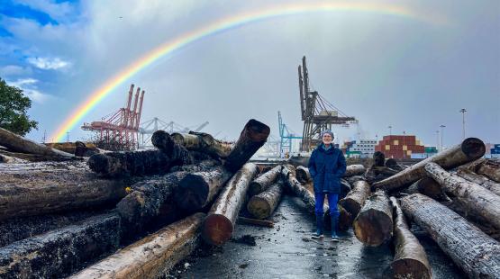 George Blomberg stands at a Port habitat restoration site among large shoreline logs, with cargo cranes and stacked shipping containers in the background. A bright rainbow arcs across the cloudy sky above the terminal.