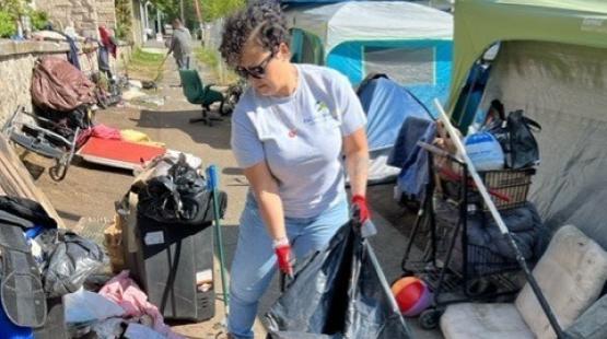 An individual lifts a large trash bag while cleaning around tents and scattered items in an outdoor encampment.