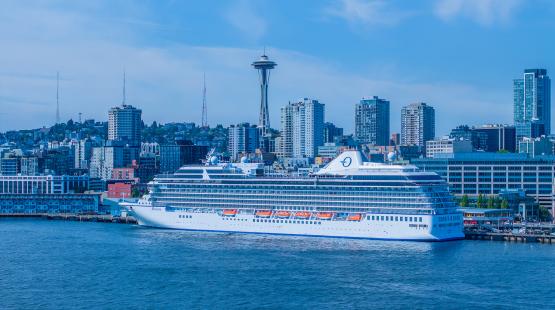 The cruise ship Oceania Riviera departing from Pier 66 in Seattle with the city skyline and the Space Needle in the background.
