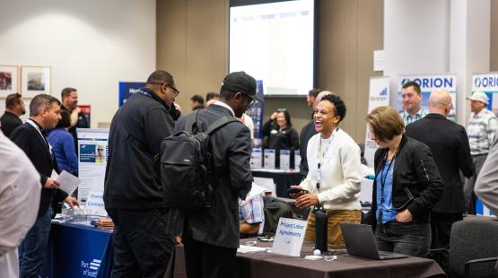People gather and talk around tables at an indoor event, including a table labeled “Project Labor Agreements” with a laptop and materials. Signs for “Diversity in Contracting” and “ORION” are visible in the background.