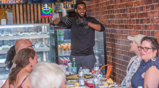 A guide at Culture Generation Food Tours in Kent speaks to a group of seated guests gathered around a table with plates of food inside a restaurant. The setting includes brick walls, a display case, and colorful menu signs.
