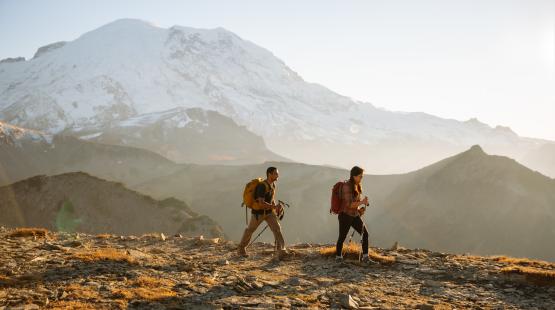 Two hikers during sunset on a trail with a snowy mountain behind them.