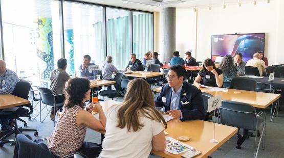 Several groups of people sit at tables in a bright meeting room, engaged in conversations. Signs with organization names, including ‘Casne’ and ‘WSP,’ are placed on some tables. Large windows with patterned columns are in the background, along with a wall‑mounted screen.