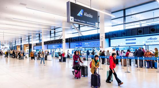 A spacious airport check‑in area with multiple travelers pulling luggage and queuing at self‑service kiosks and counters. Large overhead signs display directions for Alaska Airlines services, including first‑class check‑in and special assistance. Bright lighting and wide walkways are visible throughout the terminal.