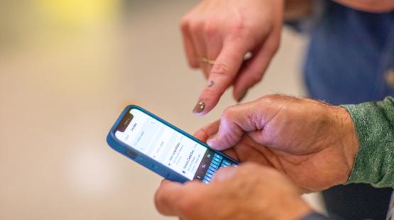 A close-up view of two hands interacting with a smartphone. One hand holds the phone while the other hand points at the screen, which displays a messaging or contacts interface. The background is blurred, placing focus on the hands and the device.