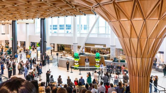 View from the Grand Stairs in the new C Concourse Expansion (CCE) building, looking down at the speaker during the CCE Grand Reveal Event. The Tree at C centerpiece of the new space is off to the right. People are seated on the amphitheater-style seating, listening to the speakers. 