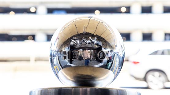 A polished metal sphere with the raised words “SAY HELLO” displayed on its front, reflecting the surrounding outdoor area including cars and people in the background.