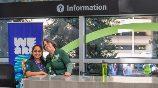 Two staff members stand behind an airport information desk. A sign above them reads ‘Information,’ and a banner beside the desk displays the words ‘We Are.’ A large window behind the desk shows trees and part of the terminal roadway. A green water bottle sits on the counter.