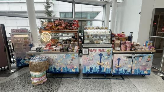 An indoor kiosk labeled “Kawaii Treats,” featuring pastel dessert illustrations on its panels. The kiosk displays shelves of packaged snacks, a refrigerated pastry case, a cold‑drink fridge, and a basket of bottled water. A small neon “Coffee & Tea” sign sits near the register. The kiosk is set inside a modern glass‑walled building with tall windows and metal beams in the background.