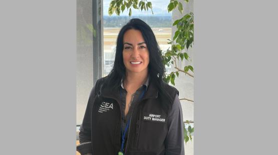 Rachel Sanders, an airport duty manager wearing a SEA Seattle‑Tacoma International Airport vest, stands indoors near a window with the airfield visible in the background and a leafy plant to the side.