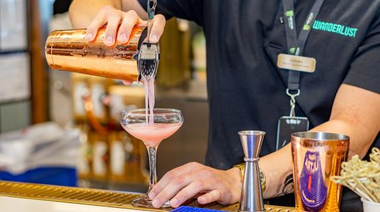 A person wearing a dark shirt with a visible name badge is pouring a pink beverage from a copper-colored cocktail shaker through a strainer into a coupe glass on a bar counter. A metal jigger and another copper-colored shaker are on the counter, with blurred bar shelves and equipment in the background.
