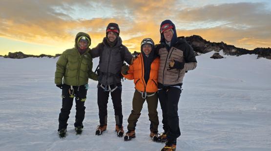 Four mountaineers in cold-weather climbing gear stand together on a snow-covered glacier at sunrise, smiling with a rocky volcanic ridge behind them.