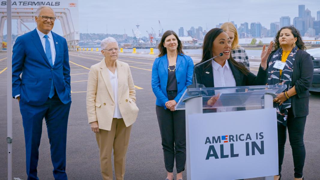 Commissioner Hasegawa speaking at an outdoor event speaking at a podium labeled 'America is all in'