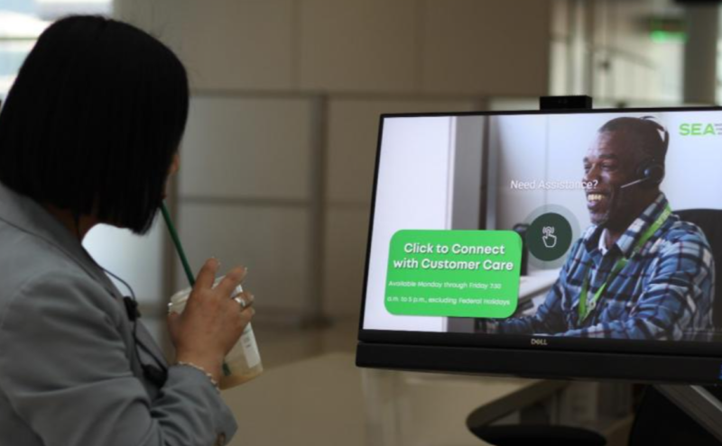 A customer in the terminal looking at a display screen that reads 'click to connect with Customer Care' and shows a happy customer service representative. 
